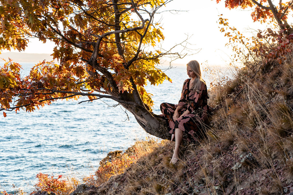A serene scene unfolds as a woman in a flowing, elegant dress sits in quiet contemplation on a naturally curved tree branch emerging from the rugged side of a mountain. The tree, artfully bent into a perfect seat, cradles her as she gazes toward the distant horizon, where the vast, shimmering expanse of Superior Lake stretches out.